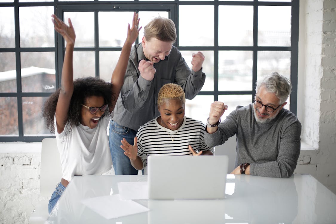 Cheerful multiethnic coworkers of different ages in casual clothing clenching fists and raising hands up in joy during successful teamwork in room